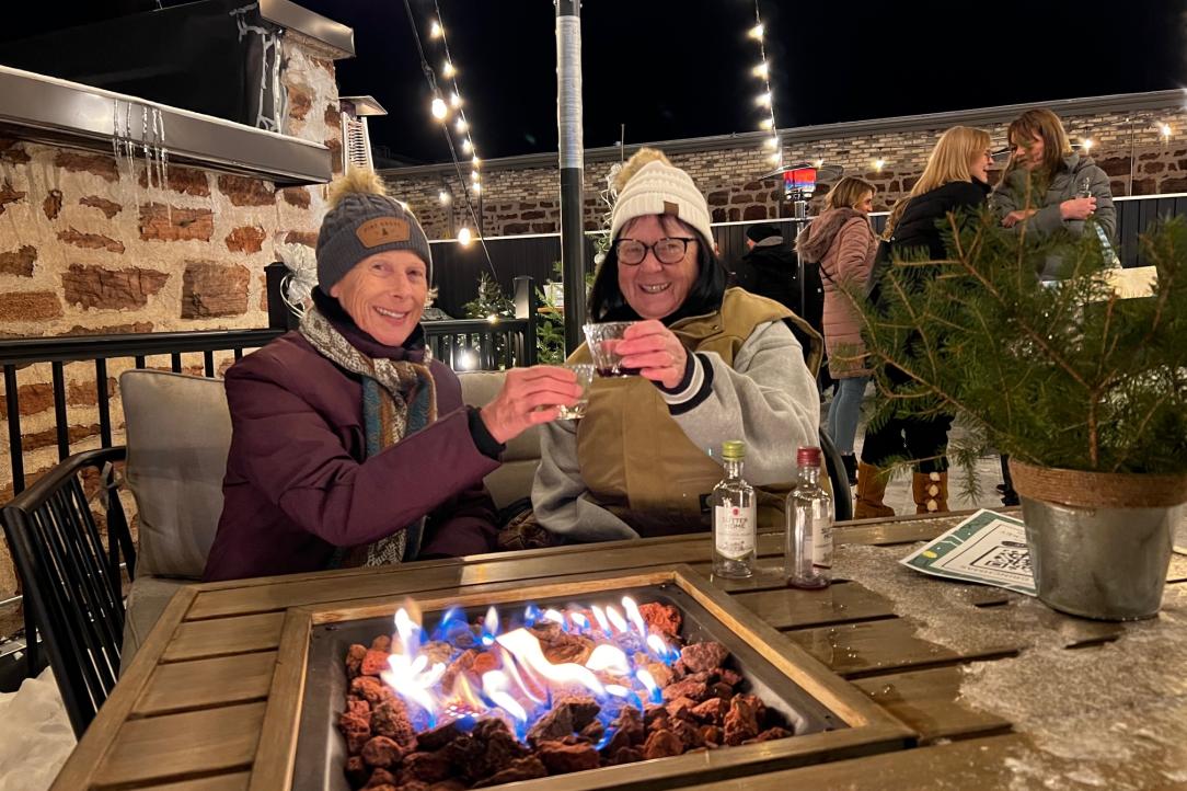 Two friends smiling at the camera with drinks near a fire